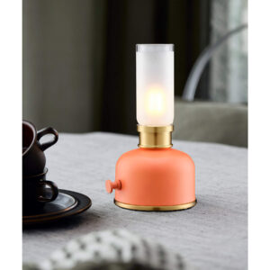 Orange bordslampa med mässingsdetaljer på grå duk, intill mörka kaffekoppar i porslin.