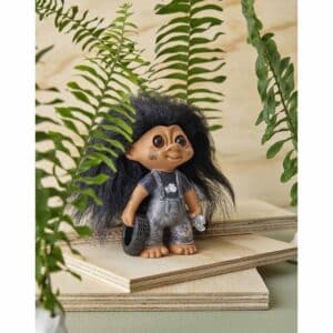 Spring Copenhagen Lyckotroll Mekaniker med svart hår och grå overall, stående bland gröna ormbunksblad på träytor.