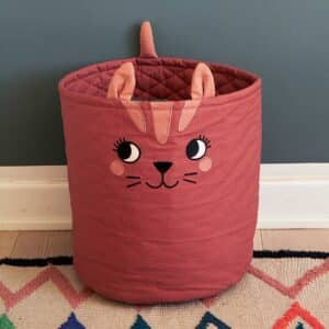 Roommate förvaringskorg i form av rostbrun katt med öron och broderat ansikte, placerad mot mörk vägg på färgglad matta.